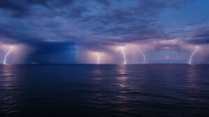 Nighttime ocean storm with several bright lightning bolts flashing across a dark, purple-tinged sky over the water.