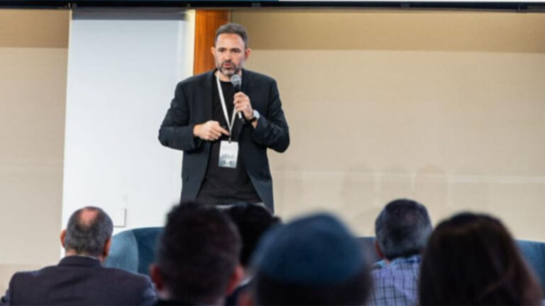 Speaker on stage presenting to an audience, holding a microphone with a lanyard badge.